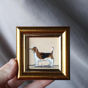 May include: A miniature framed painting of a beagle dog. The dog is shown in profile, with brown, black, and white markings. The frame is gold-coloured and square. The artwork is held by a hand.