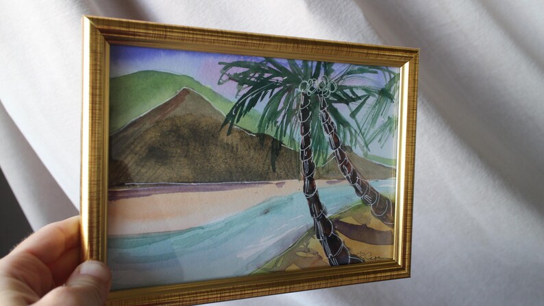May include: A framed watercolor painting of a tropical scene with two palm trees in the foreground and a mountain in the background. The painting is done in shades of blue, green, and brown.