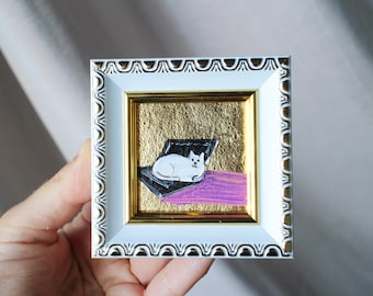 May include: A painting of a white cat in a white frame with gold accents. The cat is lying on a black laptop with a pink background.