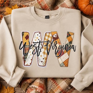 May include: A beige crewneck sweatshirt featuring a large, stylized letter W filled with a patchwork pattern of autumn-themed fabrics in shades of orange, yellow, and brown. The text West Virginia is printed in black cursive script across the letter.