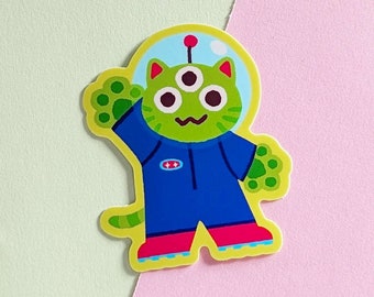Puede incluir: Un gato extraterrestre verde que lleva un traje espacial azul con una estrella amarilla en el pecho. El gato tiene tres ojos y está saludando con una pata. El gato lleva botas rojas y está de pie sobre un fondo amarillo.
