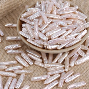 May include: A bowl filled with a large quantity of off-white, iridescent, elongated pearl beads. The beads are scattered on a beige hessian surface.