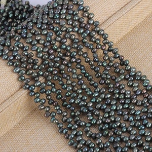 Könnte beinhalten: Stränge dunkelgrüner Süßwasserperlen. Die Perlen haben eine unregelmäßige Form und ein glänzendes Finish. Sie sind aufgefädelt und erzeugen einen kaskadierenden Effekt. Der Hintergrund ist ein neutraler beigefarbener Stoff.