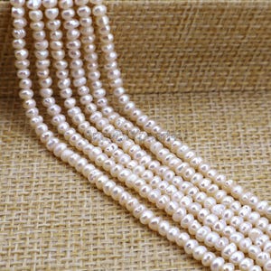 May include: Multiple strands of small, off-white pearls. The pearls are strung together, creating a layered effect. The pearls have a slightly irregular shape and a subtle lustre. The background is a textured, tan fabric.