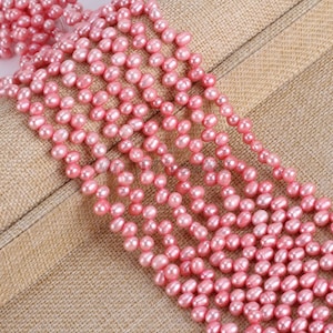 Pode incluir: Close-up de vários fios de pérolas de água doce rosa. As pérolas são ovais e têm um brilho macio e lustroso. Os fios estão dispostos em cascata, mostrando a cor e a textura das contas. O texto "AndyDIYpearl4U" é visível.