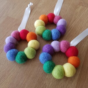 Op de afbeelding: Drie regenboog vilten bal kransen met wit lint om op te hangen. Elke krans is gemaakt van kleurrijke vilten ballen in een regenboogpatroon.