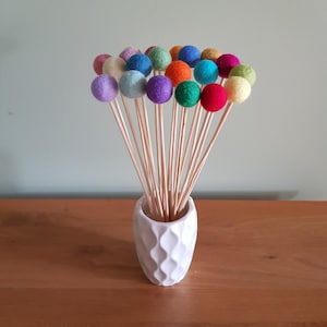 May include: A white ceramic vase with a textured surface holds a bouquet of colorful felt balls on wooden sticks. The felt balls are in various shades of blue, green, red, yellow, and purple.
