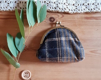 May include: A small, black and brown plaid coin purse with a metal clasp. The purse is sitting on a wooden surface with a green leafy branch and a wooden button with the text "Evelina Buttons" on it.