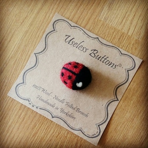 May include: A red and black needle felted ladybug brooch with a white heart on its back. The brooch is handmade in Yorkshire and is made of 100% wool. The brooch is displayed on a card with the text "Useless Buttons."