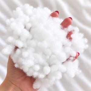 May include: A handful of white polyester fiberfill stuffing, consisting of small, rounded, cloud-like clusters of synthetic material. The texture is soft, airy, and voluminous, with a bright white color throughout the entire mass of material.