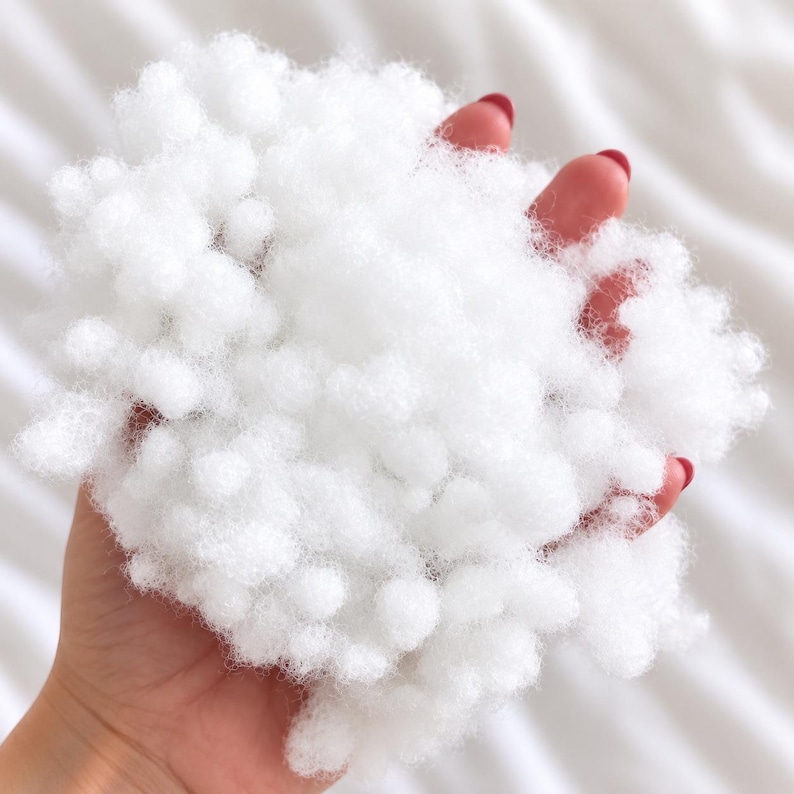 May include: A handful of white polyester fiberfill stuffing, consisting of small, rounded, cloud-like clusters of synthetic material. The texture is soft, airy, and voluminous, with a bright white color throughout the entire mass of material.