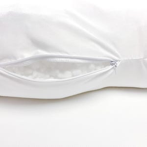 Pode incluir: Uma fronha retangular branca com um fecho de correr horizontal na lateral. O fecho est&aacute; parcialmente aberto, revelando um vislumbre do material de enchimento branco, texturizado e macio contido no interior da almofada.