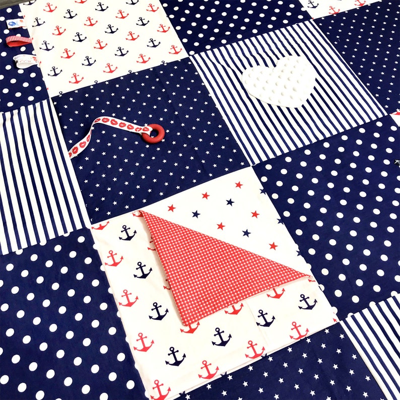 May include: A blue and white baby play mat with different textures and patterns. The mat features a white heart, red and white gingham fabric, and a blue and white striped fabric. The mat is perfect for babies to explore and learn.