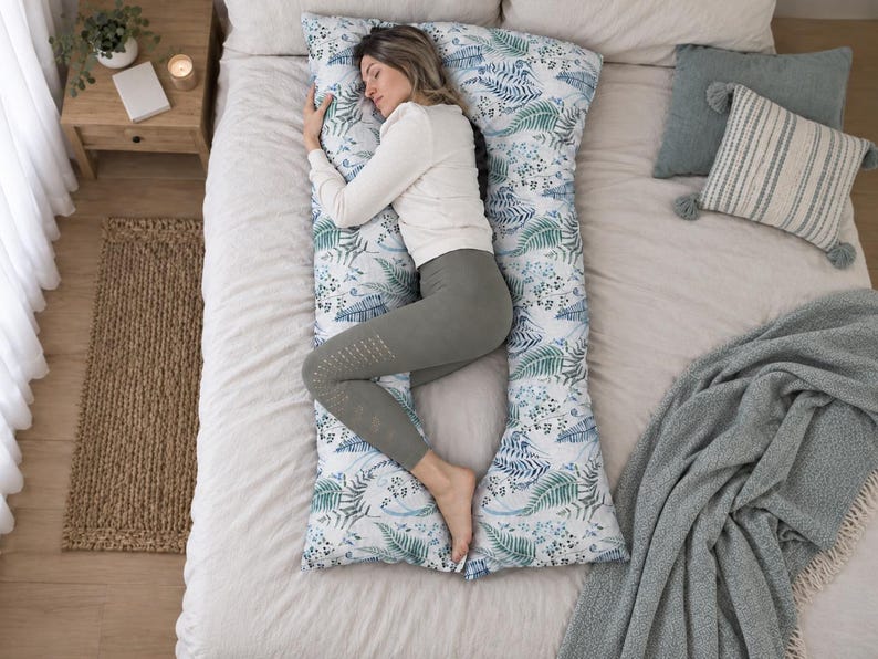 May include: A U-shaped body pillow featuring a white fabric base with a repeating botanical print of blue and green fern leaves and small dark berries. The pillow is designed to support the head, back, and legs, providing full-body cushioning.