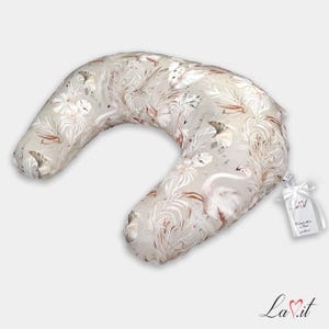 May include: A crescent-shaped nursing pillow featuring a beige fabric with a repeating pattern of white and light brown flamingos and tropical palm leaves. A white ribbon tag with the text La.it is attached to the side of the pillow.