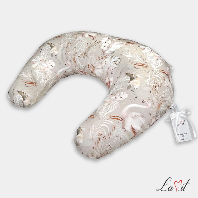 May include: A crescent-shaped nursing pillow featuring a beige fabric with a repeating pattern of white and light brown flamingos and tropical palm leaves. A white ribbon tag with the text La.it is attached to the side of the pillow.