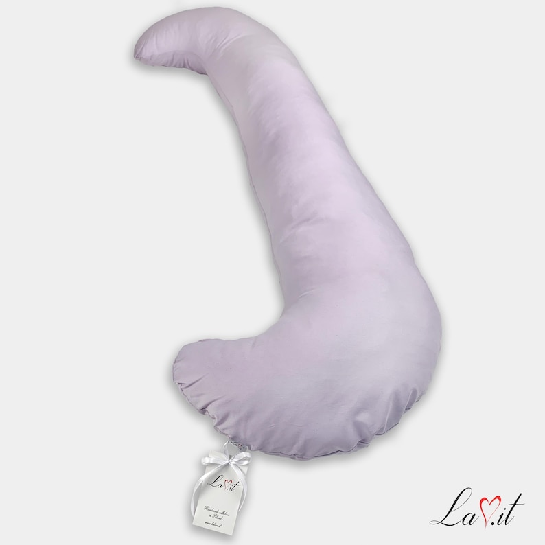 Pode incluir: Uma almofada de corpo em forma de J, de cor lavanda claro, com uma capa de tecido liso. Uma pequena etiqueta retangular branca est&aacute; presa &agrave; curva inferior com uma fita branca, apresentando o texto La.it e outros pequenos detalhes impressos.