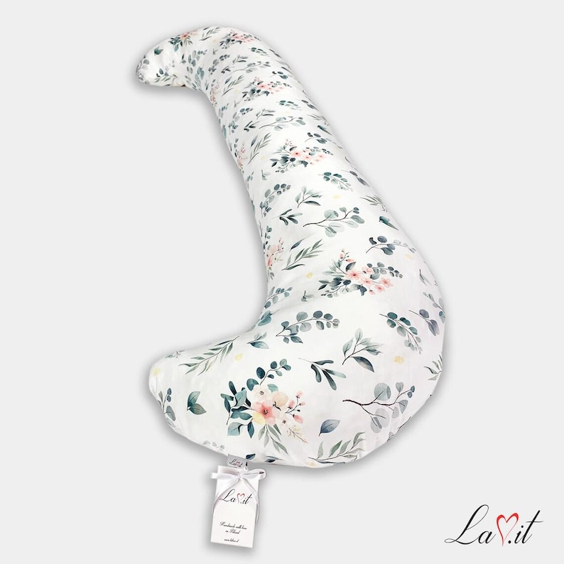 Pode incluir: Uma almofada de corpo em forma de meia-lua com uma base de tecido branco e um padr&atilde;o repetitivo de folhas de eucalipto verdes e cachos de flores cor-de-rosa. Uma pequena etiqueta retangular branca com o texto La.it est&aacute; presa na borda.