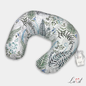 May include: A crescent-shaped nursing pillow featuring a white fabric base with a botanical print of green and dark gray ferns, blue sprigs, and light blue curved lines. A white tag with the text La.it is attached with a white ribbon.