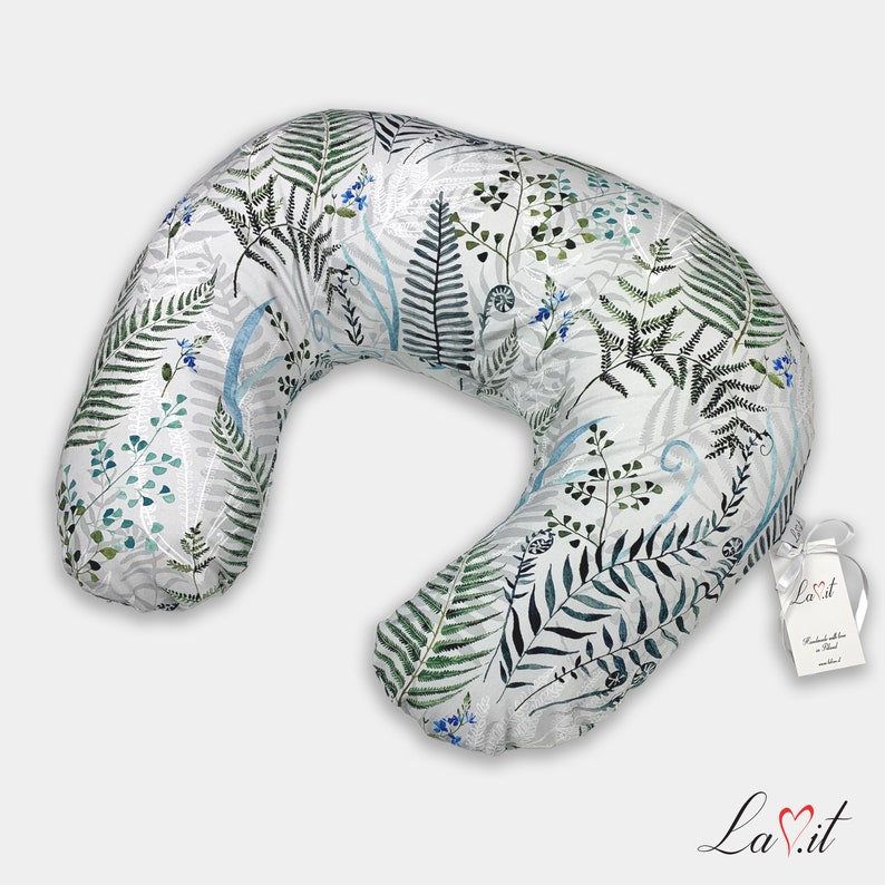 May include: A crescent-shaped nursing pillow featuring a white fabric base with a botanical print of green and dark gray ferns, blue sprigs, and light blue curved lines. A white tag with the text La.it is attached with a white ribbon.
