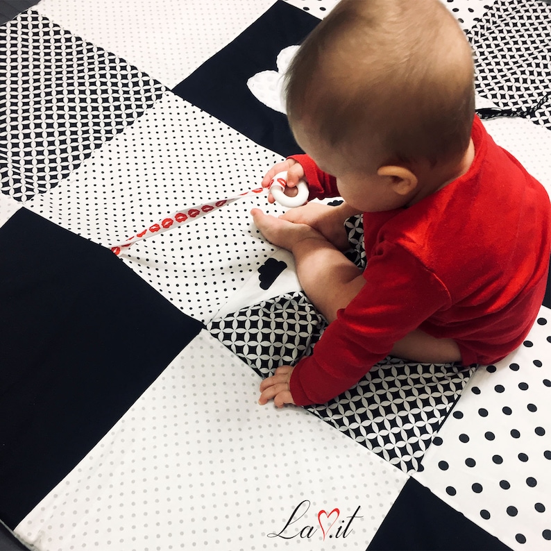 May include: A baby in a red onesie plays on a black and white patterned play mat. The mat has different textures and patterns, including polka dots, diamonds, and hearts. The baby is holding a white teething ring.
