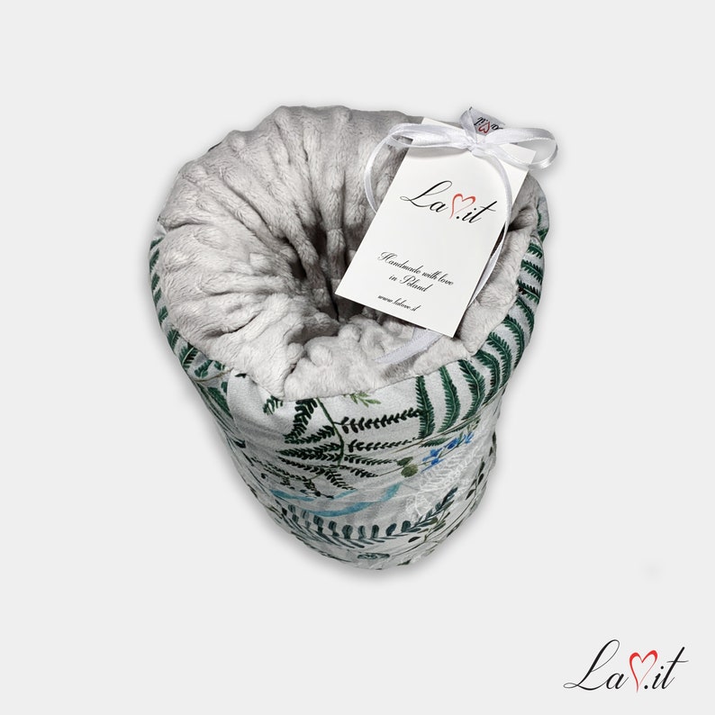 May include: A rolled blanket featuring a light grey textured interior and an exterior patterned with green fern leaves and blue floral accents. A white tag tied with a white ribbon displays the text La.it, Handmade with love in Poland, and www.lavit.pl.