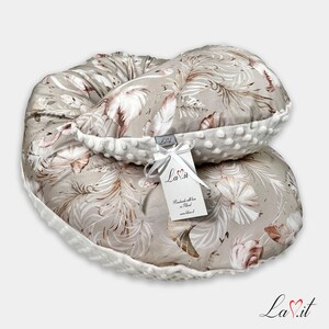 Pode incluir: Uma almofada de amamenta&ccedil;&atilde;o em forma de meia-lua com tecido bege e um padr&atilde;o repetitivo de penas brancas e castanhas. A almofada tem uma borda de tecido minky branco texturizado com pontos em relevo e uma etiqueta branca com o texto La.it.