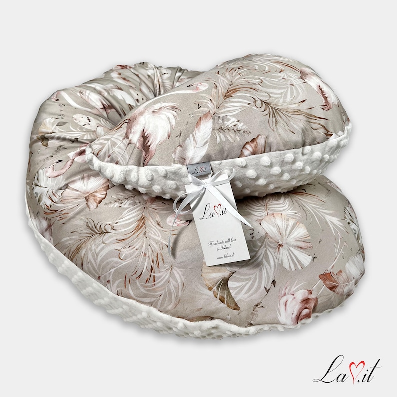 Pode incluir: Uma almofada de amamenta&ccedil;&atilde;o em forma de meia-lua com tecido bege e um padr&atilde;o repetitivo de penas brancas e castanhas. A almofada tem uma borda de tecido minky branco texturizado com pontos em relevo e uma etiqueta branca com o texto La.it.