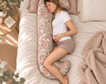 May include: A C-shaped body pillow featuring a beige background with a repeating pattern of pink flamingos and tropical foliage. The pillow is designed to support the head, back, and legs, providing a curved structure that contours to the body.