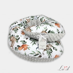 May include: A white nursing pillow set featuring a woodland animal and leaf design. The set comprises a large, circular pillow and a smaller, crescent-shaped pillow. The edges are made of grey, textured fabric. A tag reads "La Vit".