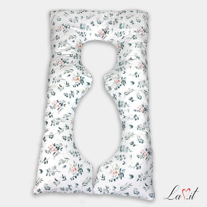 May include: A white U-shaped body pillow featuring a repeating botanical pattern of dark green eucalyptus leaves and small clusters of soft pink flowers. The pillow has a distinct contoured shape with a central cutout and the text La.it in the corner.