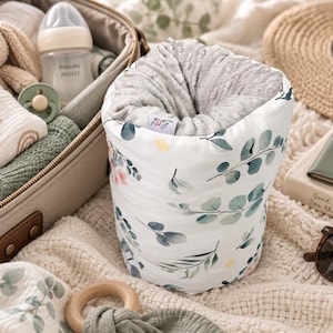 May include: A cylindrical fabric storage container featuring a white exterior with a green eucalyptus leaf and small pink flower pattern. The interior is lined with a textured grey fabric and includes a small white tag with the text Lovi.