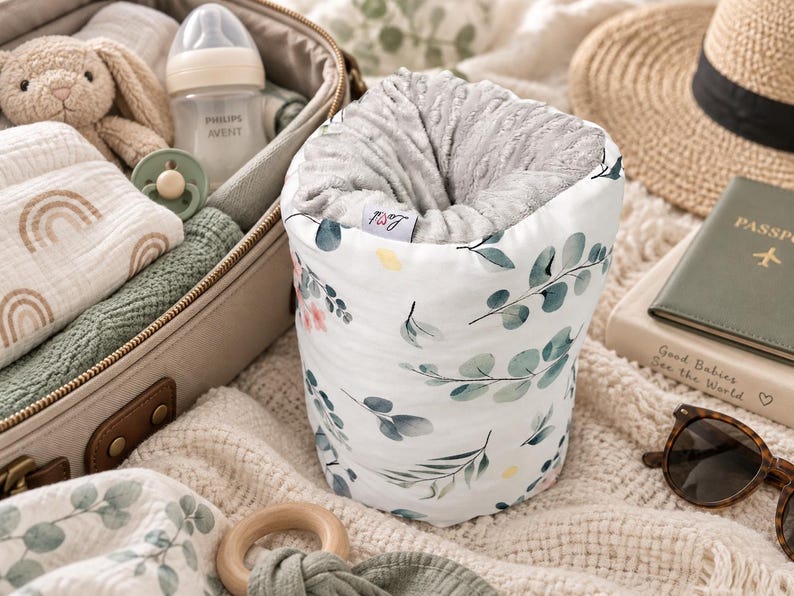 May include: A cylindrical fabric storage container featuring a white exterior with a green eucalyptus leaf and small pink flower pattern. The interior is lined with a textured grey fabric and includes a small white tag with the text Lovi.