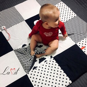 May include: A black and white baby play mat with different patterns, including polka dots, geometric shapes, and clouds. A baby is sitting on the mat, playing with a black and white patterned pillow.