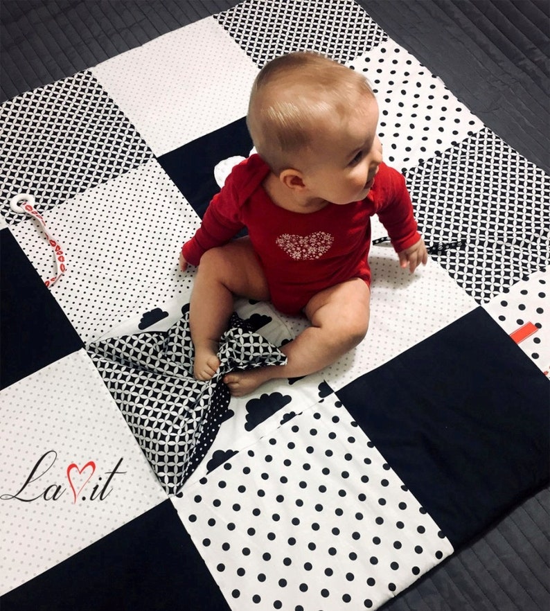 May include: A black and white baby play mat with different patterns, including polka dots, geometric shapes, and clouds. A baby is sitting on the mat, playing with a black and white patterned pillow.