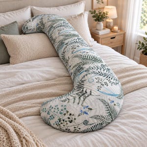 Pode incluir: Uma almofada de corpo longa e curva com uma capa de tecido branco com uma estampa botânica de folhas de samambaia verdes e pequenas flores azuis. A almofada está posicionada na diagonal sobre uma cama com roupa de cama branca e bege.
