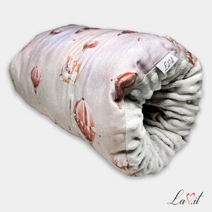 May include: A cylindrical plush pillow featuring a light pink exterior with a pattern of hot air balloons and small animals. The interior is lined with a textured grey fabric. A white fabric tag with the text La.it is attached to the side.