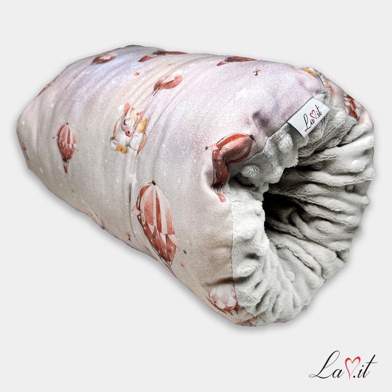 May include: A cylindrical plush pillow featuring a light pink exterior with a pattern of hot air balloons and small animals. The interior is lined with a textured grey fabric. A white fabric tag with the text La.it is attached to the side.