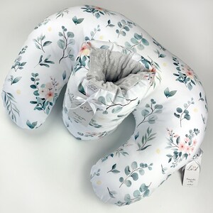 May include: A crescent-shaped nursing pillow featuring a white fabric cover with a botanical print of green eucalyptus leaves and soft pink flowers. The centre section has a grey textured lining and a white tag with the brand name Lavit.
