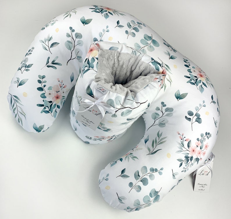 May include: A crescent-shaped nursing pillow featuring a white fabric cover with a botanical print of green eucalyptus leaves and soft pink flowers. The centre section has a grey textured lining and a white tag with the brand name Lavit.