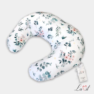 May include: A crescent-shaped nursing pillow featuring a white fabric base with a repeating watercolor pattern of green eucalyptus leaves and small pink flowers. A white rectangular tag with the text La.it is attached with a white ribbon.
