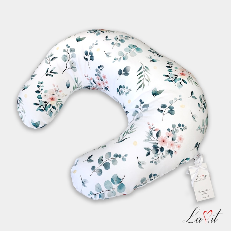 May include: A crescent-shaped nursing pillow featuring a white fabric base with a repeating watercolor pattern of green eucalyptus leaves and small pink flowers. A white rectangular tag with the text La.it is attached with a white ribbon.