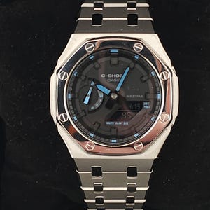 May include: A silver G-Shock Casio watch with a black face and blue detailing. The watch has a digital display showing the date and time, and a silver metal strap. The watch is presented on a black surface.