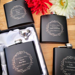 May include: Set of six black engraved hip flasks with a silver funnel. Each flask has a different name and title, such as "Best Man" and "Father of the Bride". The flasks are decorated with a floral wreath design.