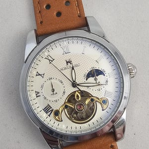 May include: A silver wristwatch with a cream face, Roman numerals, and a brown leather strap. The watch has a visible mechanical movement and the text "AOKULASIC". The strap has a series of holes.