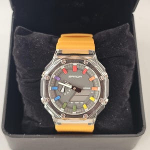Puede incluir: Un reloj digital con caja transparente y correa naranja. La esfera del reloj es gris con detalles coloridos y el texto "SANDA". El reloj se muestra en una caja negra.