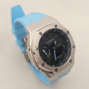 May include: A G-Shock Casio watch featuring a brushed silver octagonal metal bezel with eight visible screws, a black analog-digital dial with light blue accents, and a light blue silicone strap with a matching buckle and integrated lug design.