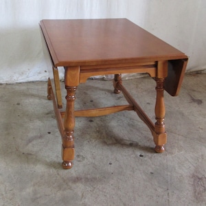 May include: A wooden drop leaf table with a brown finish. The table has four legs with turned details and a rectangular top.