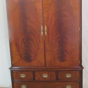 May include: A tall, wooden armoire with a rich, reddish-brown finish. The upper section features two doors with oval inlays, while the lower section has drawers with brass-colored knobs. The armoire is a classic piece of furniture.