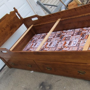 May include: A brown wooden daybed frame featuring a high headboard with turned posts, three lower storage drawers with metal pulls, and a base lined with a brown and white patterned fabric. The frame includes three horizontal wooden slats.